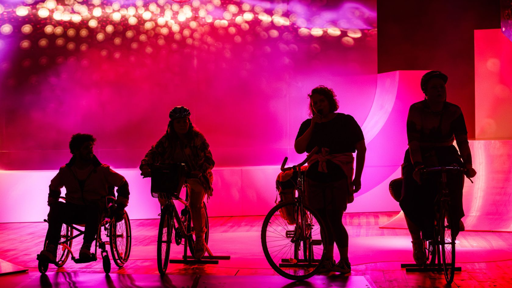cztery czarne cienie osób jadących na rowerach i na wózku inwalidzkim na różowym tle