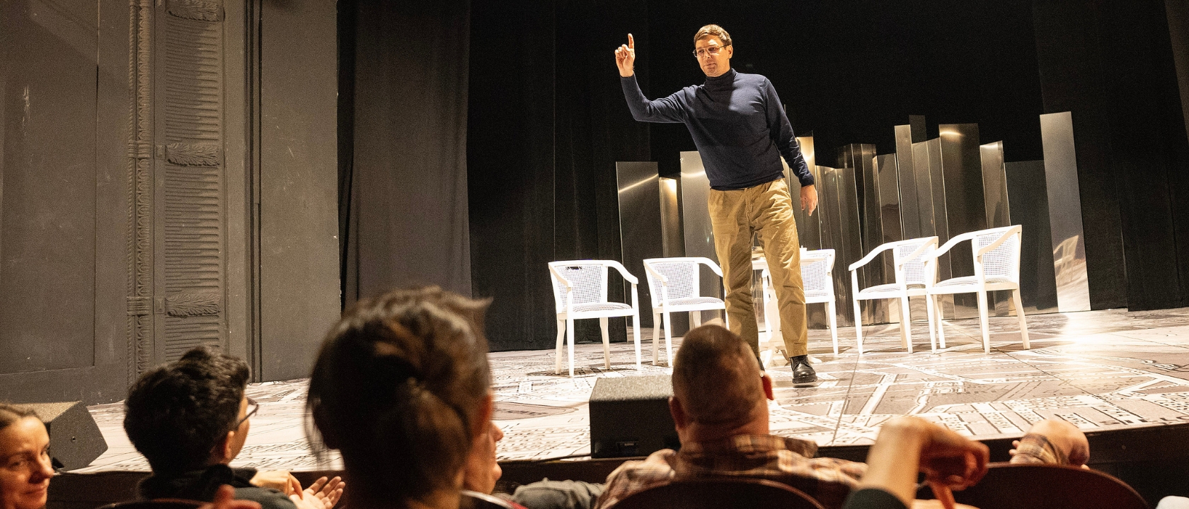 na zdjęciu mężczyzna stoi na scenie, trzyma palec uniesiony w górę, na widowni siedzą widzowie
