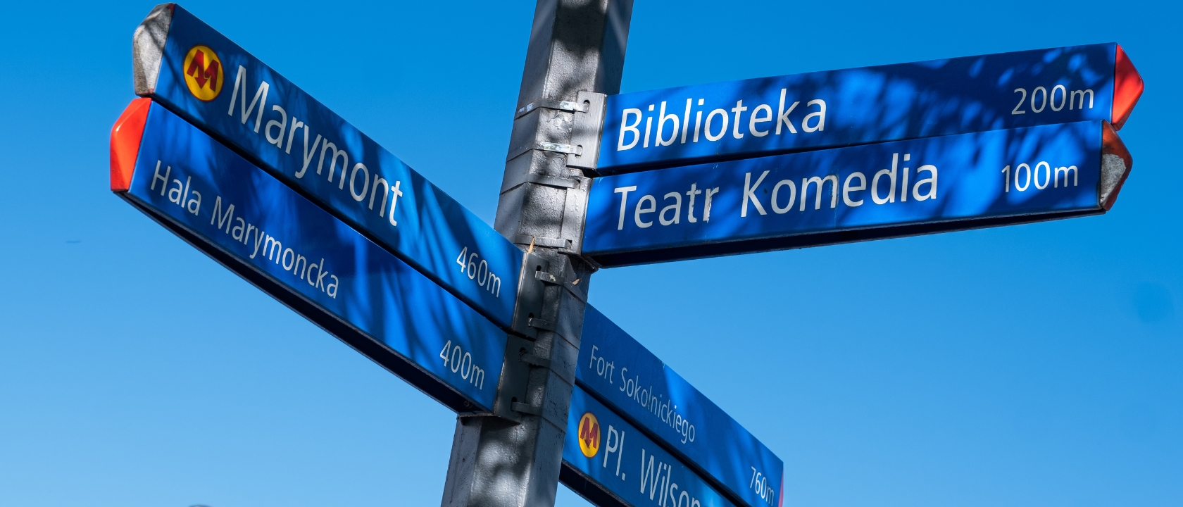 zdjęcie przedstawia słup na ulicy, który pokazuje różne kierunki - są napisane Biblioteka, Teatr Komedia, Marymont