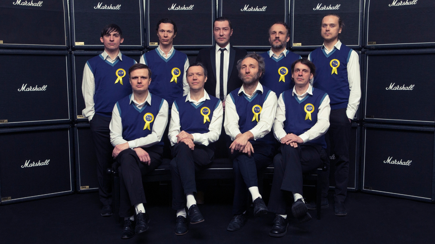 zdjęcie przedstawia zespół Mitch & Mitch - kilkoro mężczyzn w mundurkach siedzących na krzesłach i pozujących do zdjęcia na tle głośników Marshall